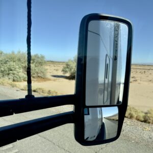 Side mirror view of an M2GT tractor-trailer reflecting the highway, with desert scrub and open sand stretching out under a clear blue sky